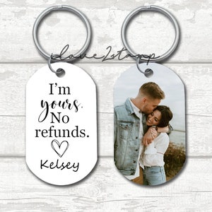 May include: A white oval keychain with the text "I'm yours. No refunds." and a heart symbol. Below the text is the name "Kelsey". The other side of the keychain features a photo of a couple embracing.
