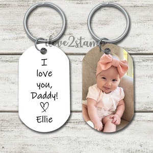 May include: Two oval-shaped keychains. One keychain has a white background with black text that reads "I love you, Daddy!" with a heart symbol and the name "Ellie". The other keychain has a photo of a baby girl wearing a pink headband and a white dress.