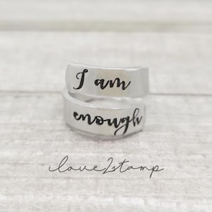 May include: A silver-colored wrap ring with the words "I am enough" in a handwritten font. The ring is on a light-colored surface. The text "ilove2stamp" is at the bottom of the image.