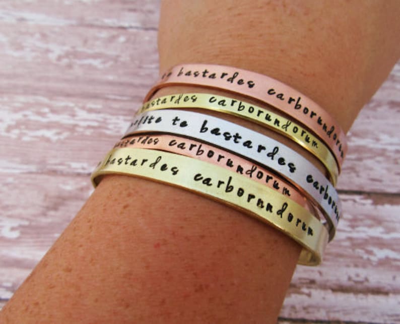 May include: A set of four stacked metal bracelets with the words "Little to Bastardes Carborundum" engraved on them. The bracelets are in different colors, including gold, silver, and copper.