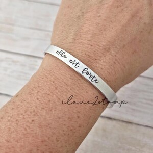 May include: A silver bracelet with the inscription "elle est forte" on a person's wrist.