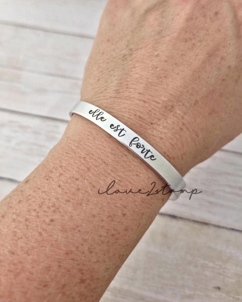 May include: A silver bracelet with the inscription "elle est forte" on a person's wrist.