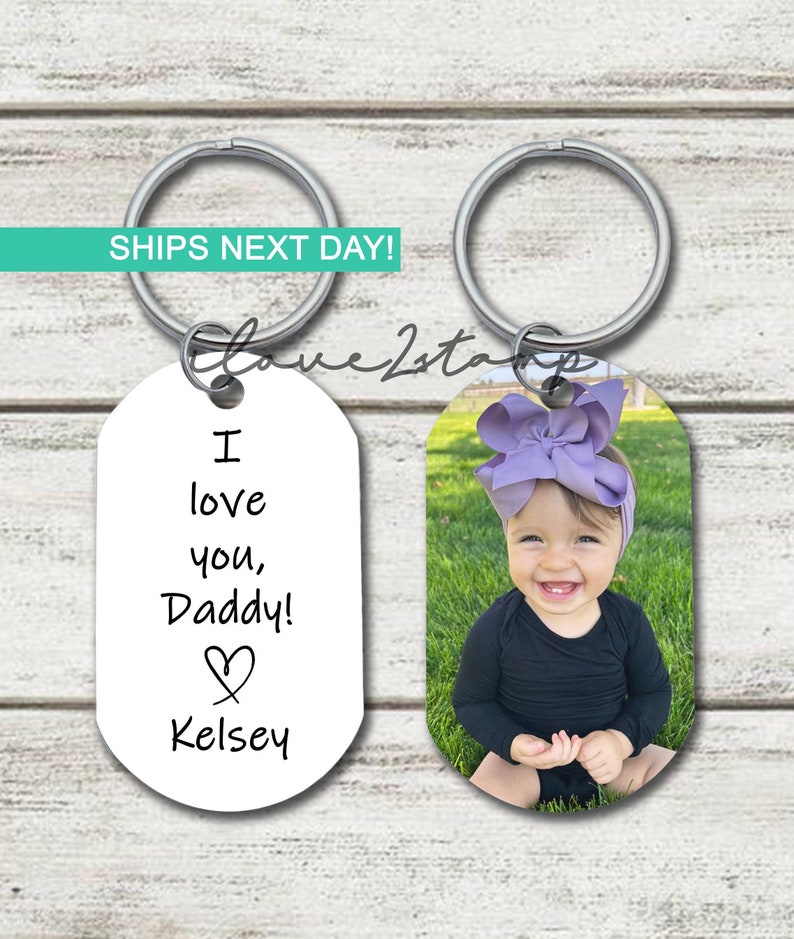 May include: Two oval-shaped keychains with silver key rings. One keychain has a white background with black text that reads "I love you, Daddy!" with a heart symbol and the name "Kelsey". The other keychain has a photo of a young girl with a purple bow in her hair, smiling, wearing a black long-sleeved shirt, sitting on green grass.