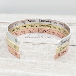 May include: Three stacked silver, gold, and rose gold bracelets with engraved text. The top bracelet reads "Once by my side, forever in my heart". The middle bracelet reads "Once by my side, forever in my heart". The bottom bracelet reads "by my side, forever in my heart". The bracelets are engraved with the names "Daisy", "Layla", and "Beau".