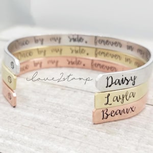 May include: Three silver, gold, and copper bracelets stacked on top of each other. The top bracelet has the inscription "Once by my side, forever in my". The middle bracelet has the inscription "Once by my side, forever in my". The bottom bracelet has the inscription "Once by my side, forever". The bottom bracelet also has the inscription "Daisy" on the side. The middle bracelet has the inscription "Layla" on the side. The bottom bracelet has the inscription "Beaux" on the side.
