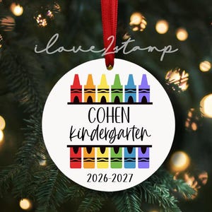Op de afbeelding: Een ronde witte kerstornament hangend aan een rood lint. Het ontwerp bevat twee rijen met kleurrijke kleurpotloodillustraties in rood, oranje, geel, groen, blauw en paars. De tekst luidt COHEN KINDERGARTEN en 2026-2027 in een zwart lettertype.