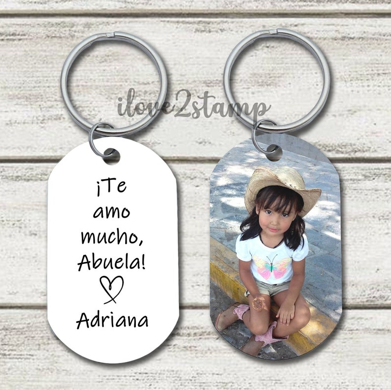 Peut inclure: Deux porte-cl&eacute;s argent&eacute;s avec une &eacute;tiquette ovale blanche. Une &eacute;tiquette a une photo d'une jeune fille portant un chapeau de paille et une chemise &agrave; imprim&eacute; papillon. L'autre &eacute;tiquette porte le texte "&iexcl;Te amo mucho, Abuela! Adriana" avec un symbole de c&oelig;ur.