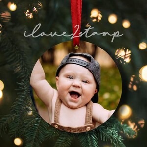 May include: A round, white, ceramic bauble with a photo of a smiling baby wearing a brown and black hat. The bauble is hanging from a red ribbon.