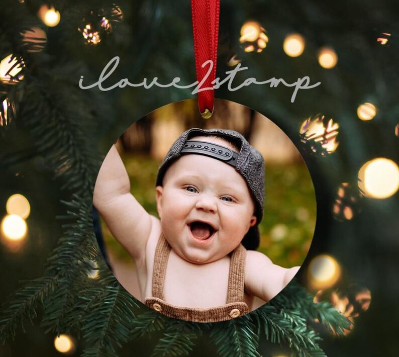 May include: A round, white, ceramic bauble with a photo of a smiling baby wearing a brown and black hat. The bauble is hanging from a red ribbon.