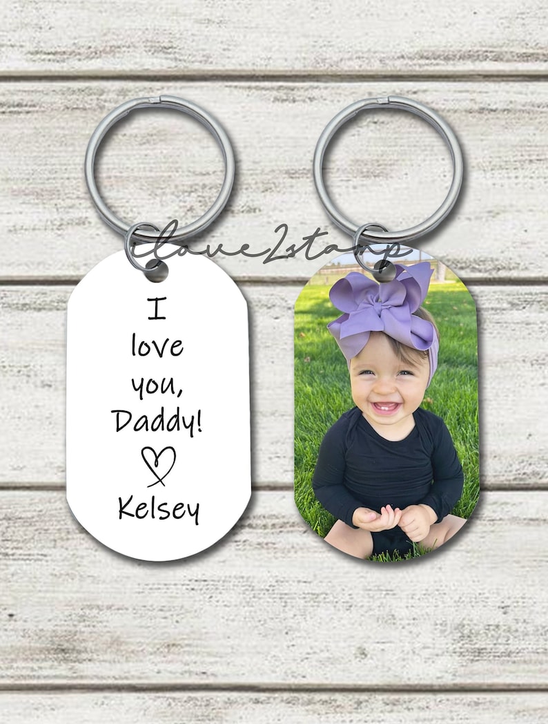 May include: Two silver keychains with oval-shaped tags. One tag has a photo of a young girl with a purple bow in her hair, smiling. The other tag has the text "I love you, Daddy!" with a heart and the name "Kelsey".