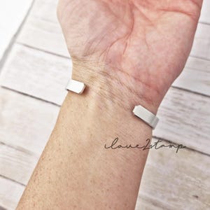 May include: A silver cuff bracelet on a person's wrist. The bracelet is open on one side and has a simple, flat design.