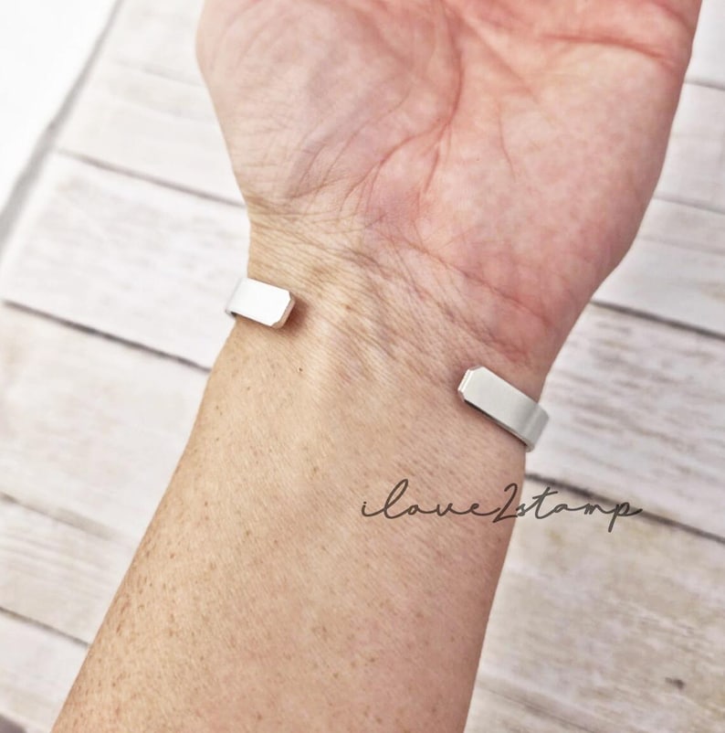 May include: A silver cuff bracelet on a person's wrist. The bracelet is open on one side and has a simple, flat design.