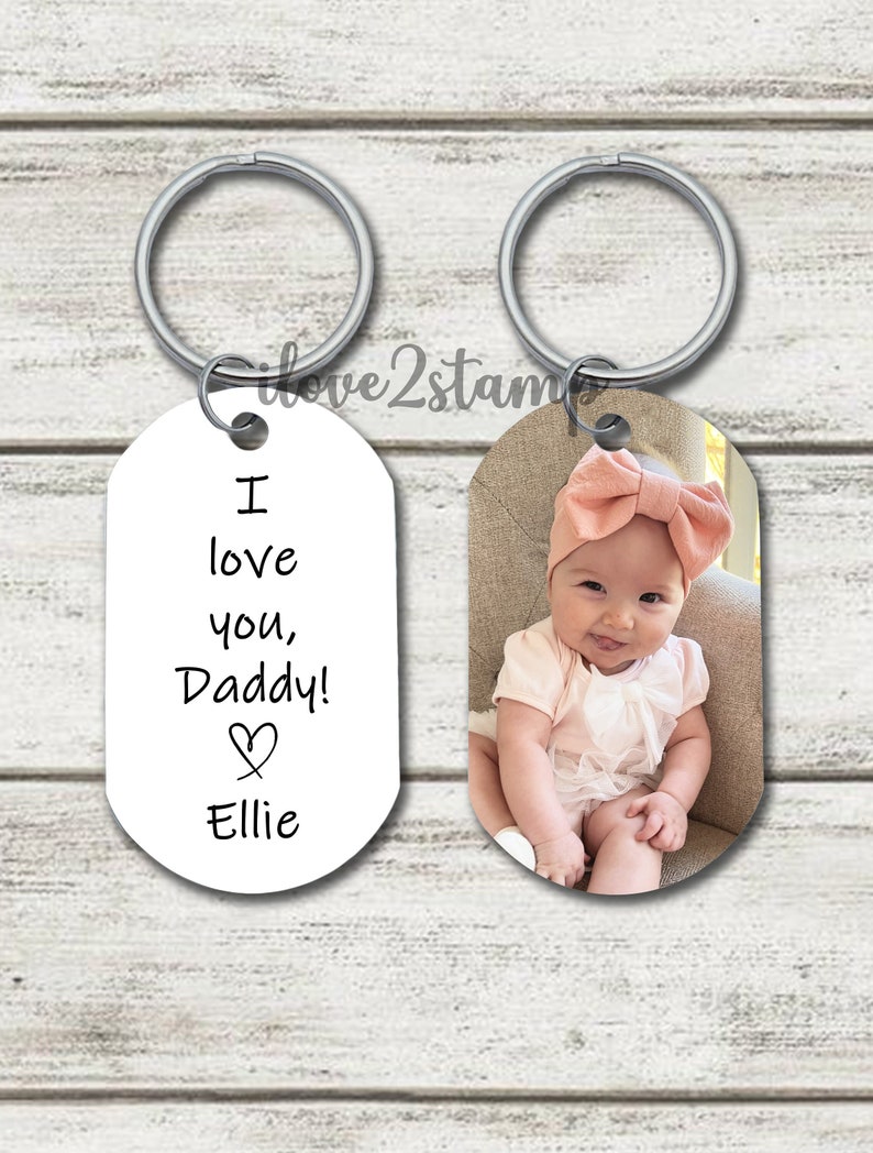 May include: Two oval-shaped keychains. One keychain has a white background with black text that reads "I love you, Daddy!" with a heart symbol and the name "Ellie". The other keychain has a photo of a baby girl wearing a pink headband and a white dress.