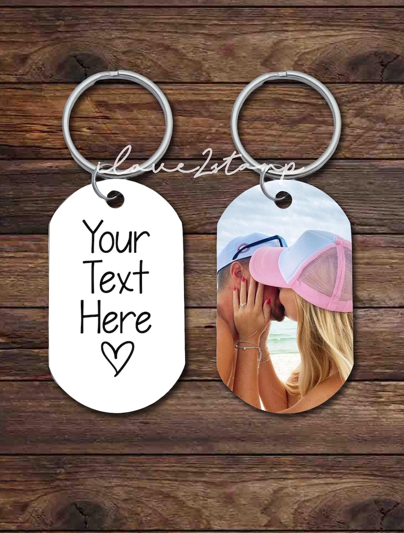 K&ouml;nnte beinhalten: Zwei silberne Schl&uuml;sselanh&auml;nger mit ovalen Anh&auml;ngern. Ein Anh&auml;nger ist wei&szlig; mit schwarzem Text, der "Your Text Here" und ein Herz sagt. Der andere Anh&auml;nger hat ein Foto von einem Paar, das sich am Strand k&uuml;sst.
