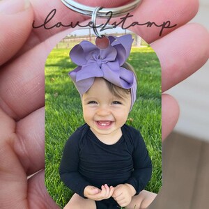 May include: A silver keyring with a photo of a baby girl wearing a purple bow and a black shirt. The baby is sitting on a green grassy lawn.