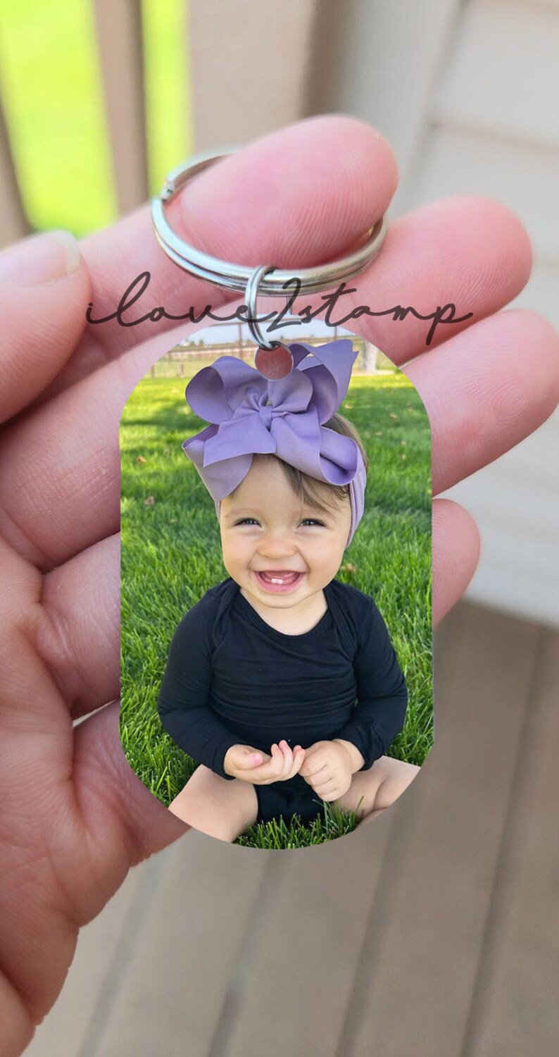 May include: A silver keyring with a photo of a baby girl wearing a purple bow and a black shirt. The baby is sitting on a green grassy lawn.