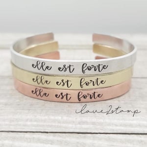 May include: Three stacked bracelets with the phrase "elle est forte" engraved on each. The bracelets are made of silver, gold, and copper.