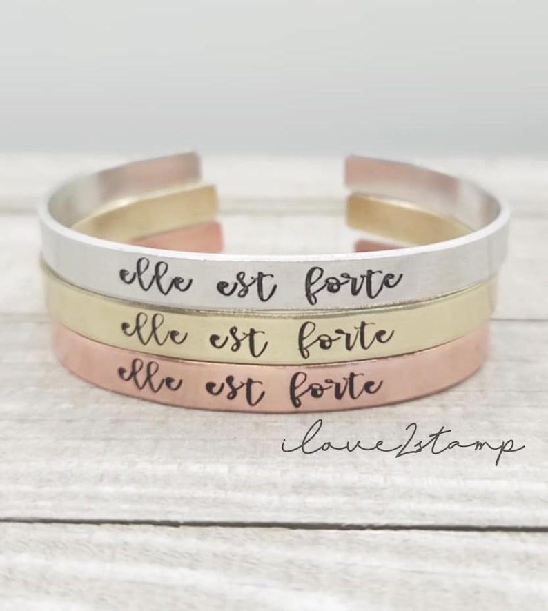 May include: Three stacked bracelets with the phrase "elle est forte" engraved on each. The bracelets are made of silver, gold, and copper.