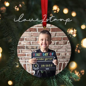 Op de afbeelding: Een ronde keramische kerstornament met een foto van een lachend kind dat een zwart bordje vasthoudt met de tekst FIRST DAY OF kindergarten, 5 YEARS OLD en 08/13/25. Het ornament hangt aan een rood lint tegen een wazige groene achtergrond.