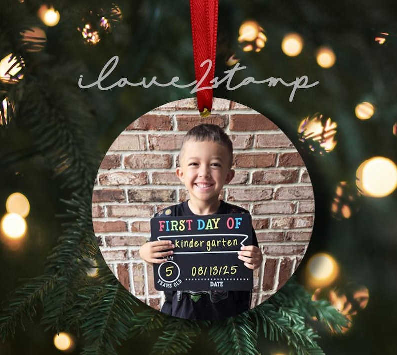 Op de afbeelding: Een ronde keramische kerstornament met een foto van een lachend kind dat een zwart bordje vasthoudt met de tekst FIRST DAY OF kindergarten, 5 YEARS OLD en 08/13/25. Het ornament hangt aan een rood lint tegen een wazige groene achtergrond.