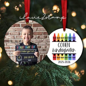 Op de afbeelding: Twee ronde keramische ornamenten met rode linten. E&eacute;n ornament toont een foto van een kind met een schoolbordje met de tekst FIRST DAY OF kindergarten 5 YEARS OLD 08/13/25. De andere toont kleurrijke kleurpotloodafbeeldingen, de naam COHEN, de tekst kindergarten en 2025-2026.
