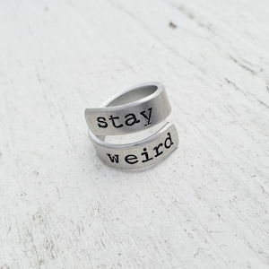 Może przedstawiać: Srebrny pierścionek z wygrawerowanym napisem "stay weird". Pierścionek jest wykonany z metalu i ma prosty design.
