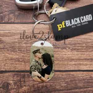 以下が含まれることがあります： 黒と黄色のカードに「pf BLACK CARD ALL THE PERKS」と書かれたシルバーのキーホルダーと、カップルが写った小さな楕円形のフォトチャーム。男性は軍服を着ており、女性は黒いシャツを着ています。