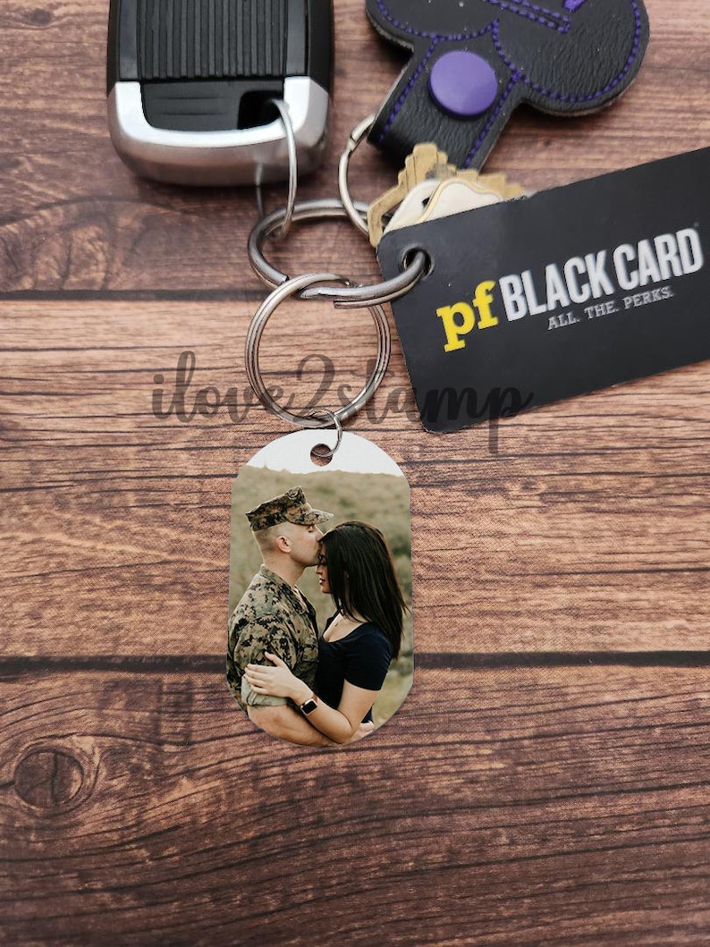 以下が含まれることがあります： 黒と黄色のカードに「pf BLACK CARD ALL THE PERKS」と書かれたシルバーのキーホルダーと、カップルが写った小さな楕円形のフォトチャーム。男性は軍服を着ており、女性は黒いシャツを着ています。