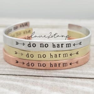 Puede incluir: Tres pulseras apiladas, cada una grabada con la frase "no hagas daño". Las pulseras están hechas de diferentes metales: plata, oro y cobre.