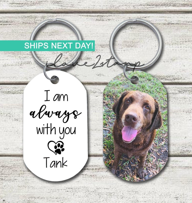 May include: A white oval keyring with the text "I am always with you" and a paw print heart. The text "Tank" is below the heart. A second oval keyring features a photo of a brown dog with a pink tongue.