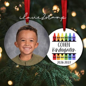 Op de afbeelding: Twee ronde ornamenten met rode linten. E&eacute;n ornament toont een foto van een lachend kind in een groen shirt. Het andere witte ornament bevat kleurrijke kleurpotloodafbeeldingen, de tekst Cohen Kindergarten en het schooljaar 2026-2027.