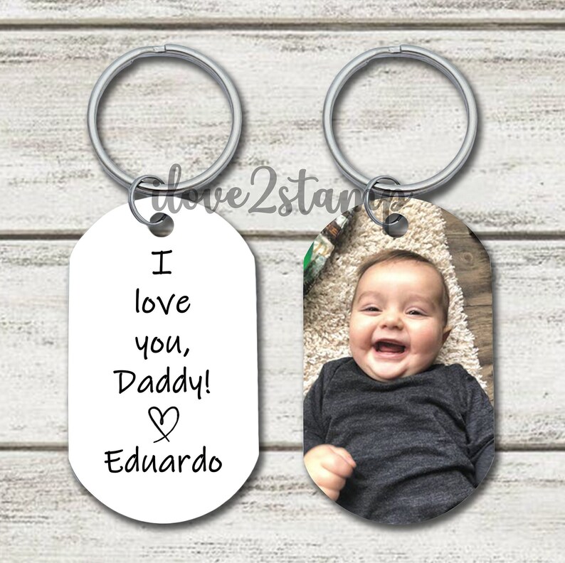 May include: A silver keychain with two oval-shaped tags. One tag has a photo of a baby boy smiling. The other tag has the text "I love you, Daddy!" with a heart symbol and the name "Eduardo".