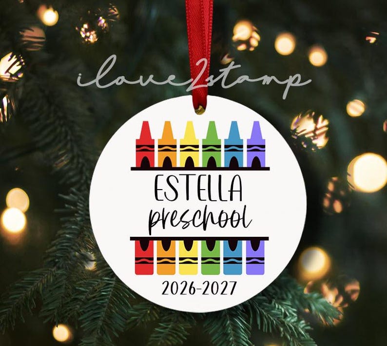 Op de afbeelding: Een rond wit keramieken ornament hangend aan een rood lint. Het ontwerp toont twee rijen kleurrijke kleurpotloodillustraties in rood, oranje, geel, groen, blauw en paars. De tekst luidt Estella preschool en 2026-2027 in een zwart lettertype.