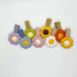 Pode incluir: Um conjunto de oito presilhas de cabelo de flores de crochê em várias cores, incluindo amarelo, laranja, marrom, branco e rosa. Cada flor está presa a uma presilha coberta de tecido.