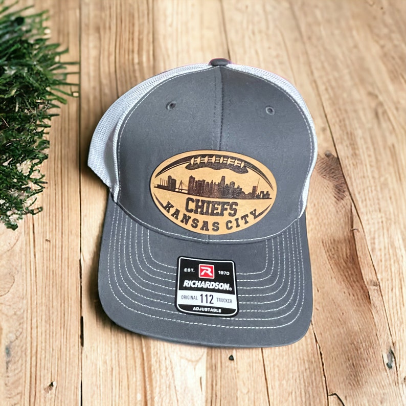 Pu&ograve; includere: Cappellino da camionista grigio e bianco con una toppa in pelle marrone che presenta uno skyline della citt&agrave; e il testo "Chiefs Kansas City". La toppa ha la forma di un pallone da calcio. Il cappello ha un cinturino regolabile nero con un logo rosso e bianco che dice "Richardson 112".