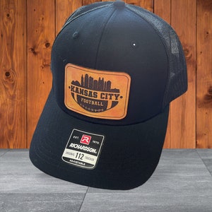 May include: Black trucker hat with a brown leather patch featuring a silhouette of the Kansas City skyline and the text "Kansas City. Football."