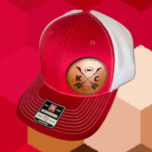 May include: Red and white trucker hat with a leather patch. The patch features crossed arrows, a football, and the letters "K" and "C". The hat has a curved brim and an adjustable strap. The tag reads "RICHARDSON ORIGINAL 112 TRUCKER ADJUSTABLE".