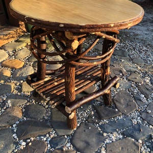 May include: A rustic wooden table with a round top and a lower shelf. The table is made of natural branches and has a light brown finish. The table is sitting on a bed of small stones.
