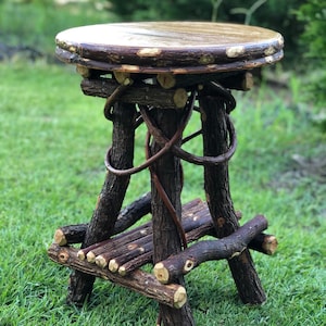 May include: A rustic wooden stool with a round top and a woven base. The stool is made of natural branches and has a dark brown finish.