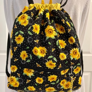 May include: Black drawstring backpack with a sunflower and bee pattern. The bag features a yellow interior, a front zipper pocket, and black shoulder straps. The bag is decorated with yellow sunflowers, bees, and white polka dots.