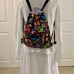 May include: A black drawstring backpack with a colorful cat print. The print features cats in various poses and colors, along with flowers and hearts. The backpack has black straps and a purple drawstring.