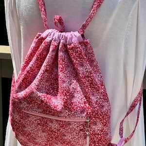 May include: A red and white floral print drawstring backpack with a zippered front pocket. The backpack has pink straps and a pink interior lining. The bag is made of fabric and is designed to be worn on the back.