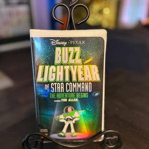May include: A Disney Pixar "Buzz Lightyear of Star Command: The Adventure Begins" VHS tape is displayed in a black metal stand. The tape features Buzz Lightyear in a green and white spacesuit. The title is in yellow and green with a space background.