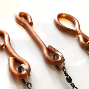 Op de afbeelding: Drie koperkleurige metalen sluitingen met een S-haakontwerp. Elke sluiting heeft aan beide uiteinden een lus, met een kleine zilveren ring en een zwarte kralenketting aan één uiteinde. De sluitingen zijn diagonaal geplaatst tegen een witte achtergrond.