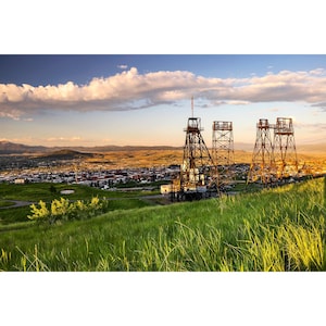 May include: A panoramic view of a small town with three rusty metal mining structures in the foreground. The structures are silhouetted against a bright blue sky with a few white clouds. The town is nestled in a valley with green hills in the background.