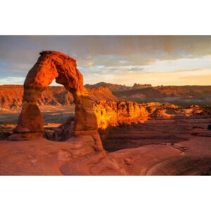 Puede incluir: Un paisaje esc&eacute;nico con un arco natural de arenisca ba&ntilde;ado por la c&aacute;lida luz del sol. El arco es de un vibrante color rojo anaranjado, que contrasta con los tonos apagados del terreno des&eacute;rtico circundante y el cielo nublado.