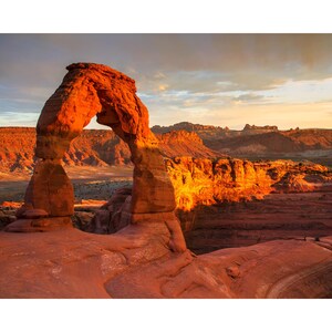 Puede incluir: Un paisaje esc&eacute;nico con un gran arco natural de arenisca ba&ntilde;ado por la luz del sol. El arco es de un vibrante color rojo anaranjado, que contrasta con el terreno des&eacute;rtico circundante y un cielo nublado. La imagen captura la belleza de una formaci&oacute;n rocosa natural.