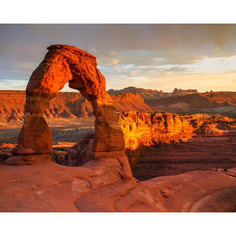Puede incluir: Un paisaje esc&eacute;nico con un gran arco natural de arenisca ba&ntilde;ado por la luz del sol. El arco es de un vibrante color rojo anaranjado, que contrasta con el terreno des&eacute;rtico circundante y un cielo nublado. La imagen captura la belleza de una formaci&oacute;n rocosa natural.