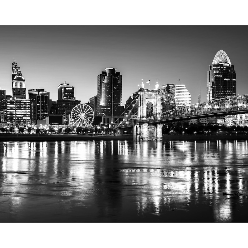 May include: A black and white photograph of a city skyline at night. The city is lit up with lights, and there is a bridge in the foreground. A Ferris wheel is visible in the distance.