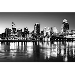 May include: A black and white photograph of the Cincinnati skyline at night. The city is lit up with lights, and a bridge crosses the river in the foreground. A Ferris wheel is visible in the distance.